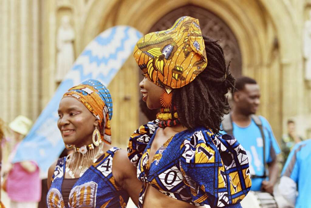 Two women in colourful wax-print headwraps and dresses smiling